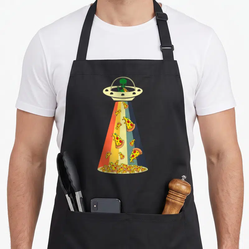 Ufo pizza apron - Green alien in beige UFO dropping multi-colored pizza slices through a rainbow striped beam — shown on a black cotton canvas apron against a white background