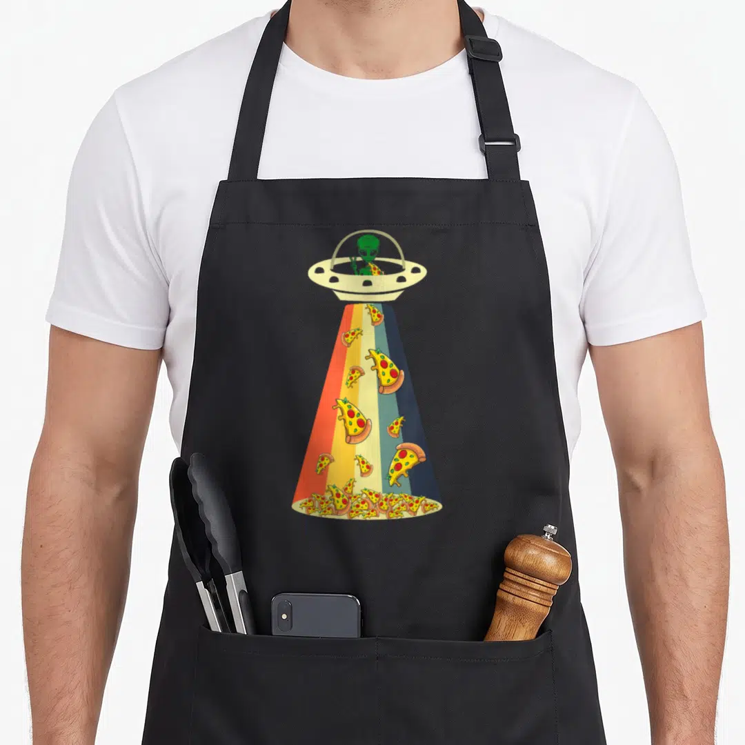 Ufo pizza apron - Green alien in beige UFO dropping multi-colored pizza slices through a rainbow striped beam — shown on a black cotton canvas apron against a white background