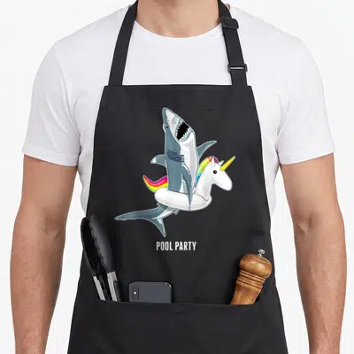 Pool unicorn apron - Cartoon shark wearing rainbow unicorn floatie with POOL PARTY text — shown on a black cotton canvas apron against a white background