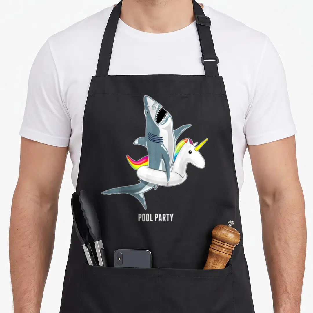 Pool unicorn apron - Cartoon shark wearing rainbow unicorn floatie with POOL PARTY text — shown on a black cotton canvas apron against a white background