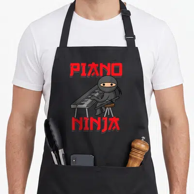 Ninja piano apron - Cartoon ninja sitting on stool playing keyboard with red PIANO NINJA text — shown on a black cotton canvas apron against a white background