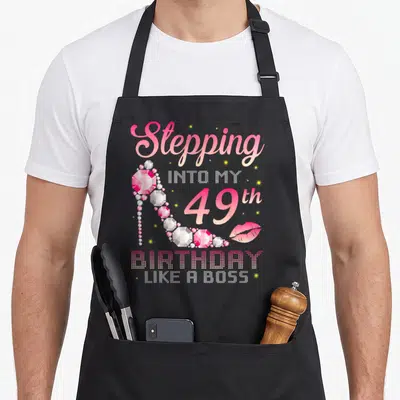 49th bbq boss apron - Pink high heel shoe surrounded by Stepping Into My 49th Birthday Like A Boss text and lips — shown on a black cotton canvas apron against a white background