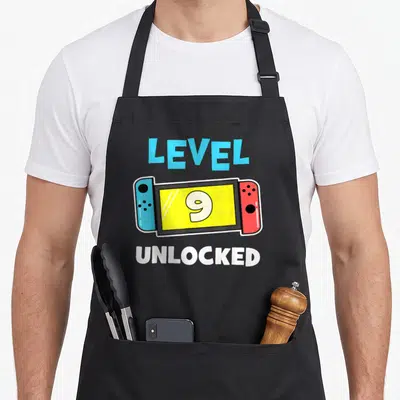 Unlocked gamer video game apron - Cartoon handheld console with blue and red controllers, yellow screen showing 9 — shown on a black cotton canvas apron against a white background