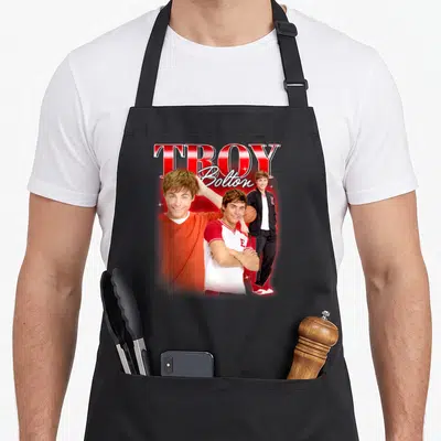 Musical school apron - Troy Bolton text with three layered photo portraits in red and orange — shown on a black cotton canvas apron against a white background