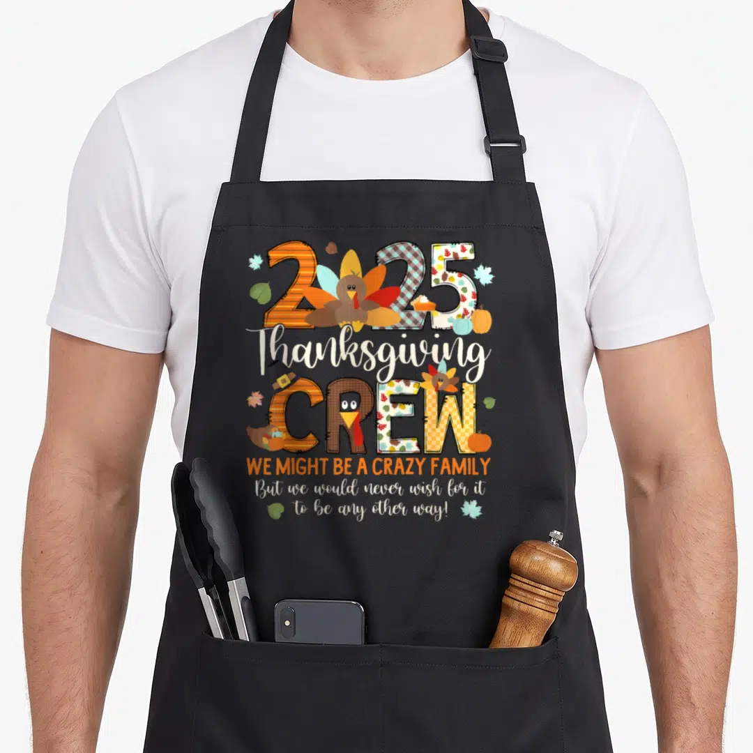 Crew thanksgiving apron - 2025 turkey cartoon with We Might Be A Crazy Family quote and autumn leaves — shown on a black cotton canvas apron against a white background