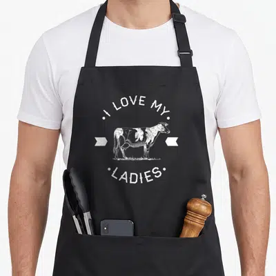 Dairy cow print aprons - White distressed cow illustration between I Love My Ladies text and two arrows — shown on a black cotton canvas apron against a white background