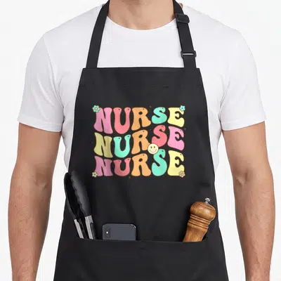 Groovy future nurse apron - Retro NURSE text in pink orange teal yellow with smiley face and flowers — shown on a black cotton canvas apron against a white background