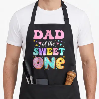 Sweet 1st donut apron - DAD of the SWEET ONE text with three colorful donuts spelling ONE surrounded by hearts and stars — shown on a black cotton canvas apron against a white background