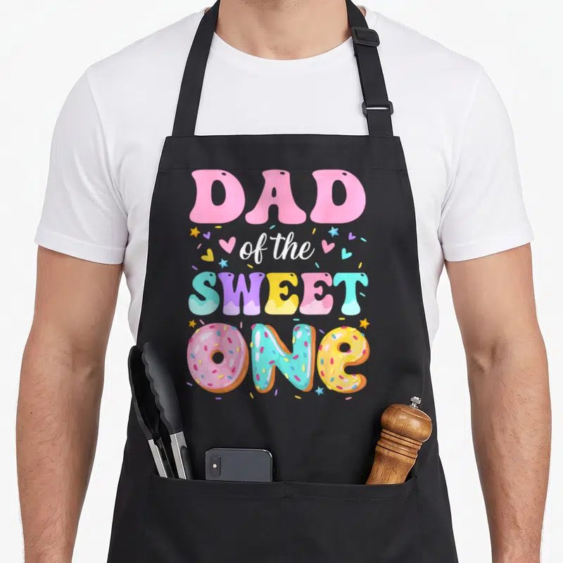 Sweet 1st donut apron - DAD of the SWEET ONE text with three colorful donuts spelling ONE surrounded by hearts and stars — shown on a black cotton canvas apron against a white background