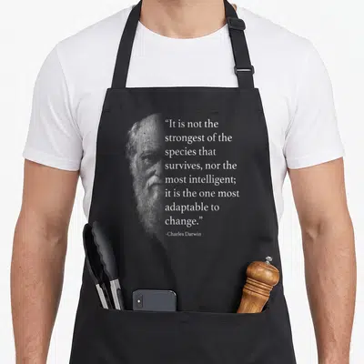 Darwin charles apron - Faded grey portrait of Charles Darwin next to white text about adapting to change — shown on a black cotton canvas apron against a white background