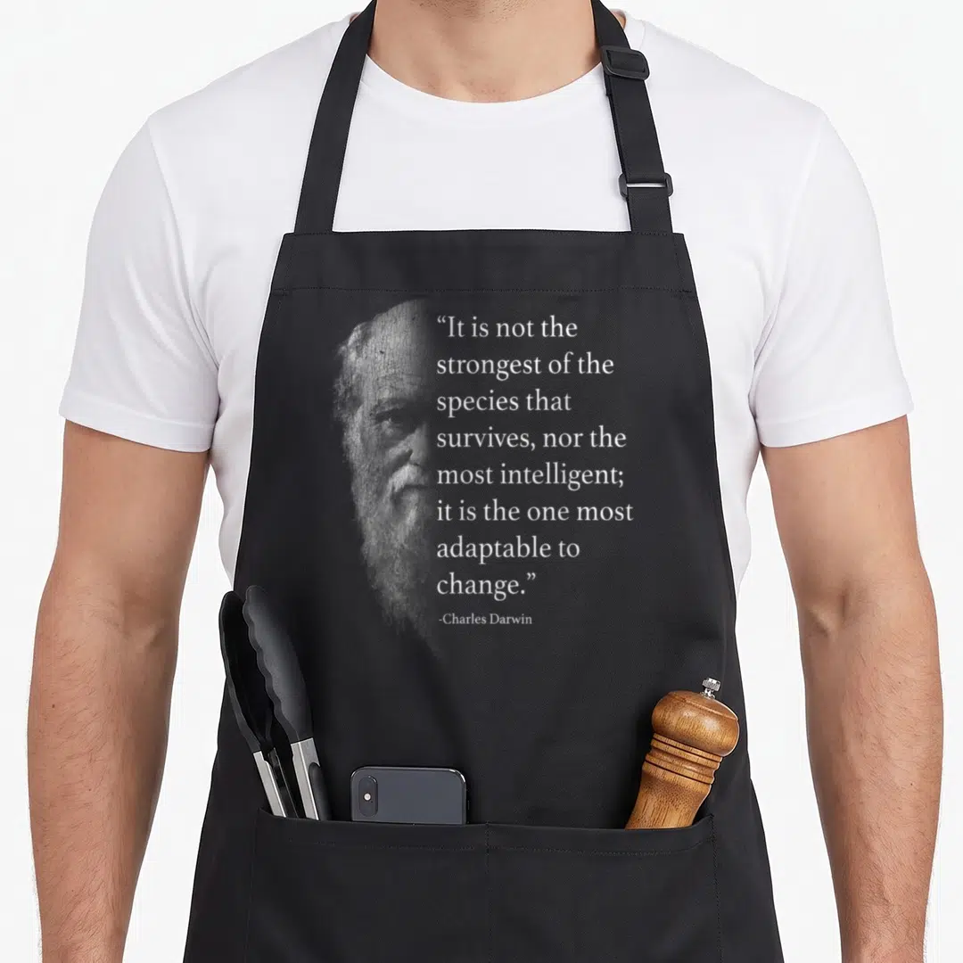 Darwin charles apron - Faded grey portrait of Charles Darwin next to white text about adapting to change — shown on a black cotton canvas apron against a white background
