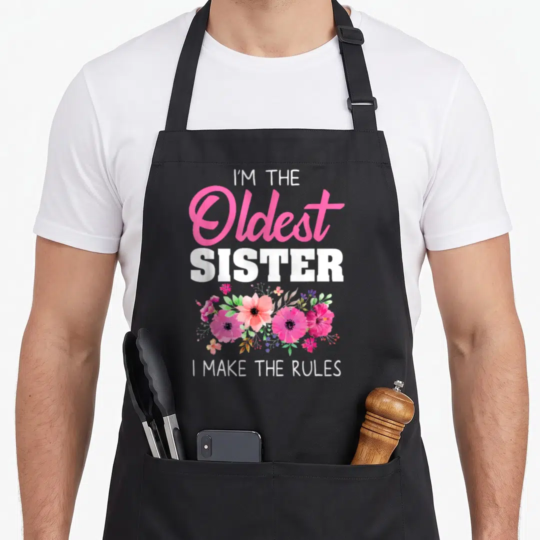 Oldest sister goals apron - I Make The Rules text with pink white and green watercolor flower cluster — shown on a black cotton canvas apron against a white background