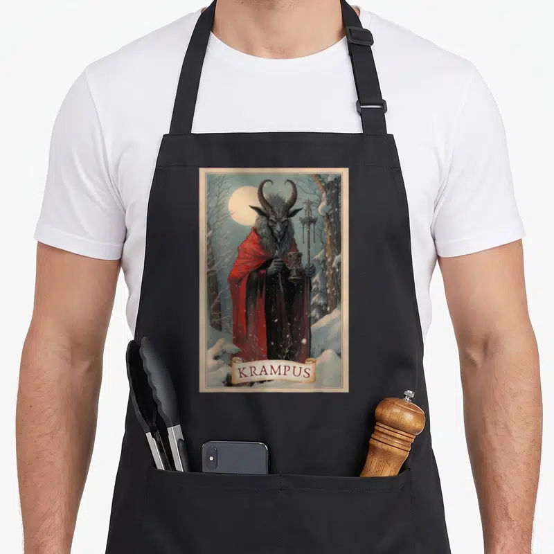 Merry xmas apron - Krampus figure in red cloak holding goblet and staff in snowy forest with banner text — shown on a black cotton canvas apron against a white background