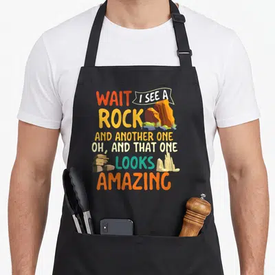 Collector hunter apron - Stacked cartoon text WAIT I SEE A ROCK with orange and teal rock illustrations on black fabric — shown on a black cotton canvas apron against a white background