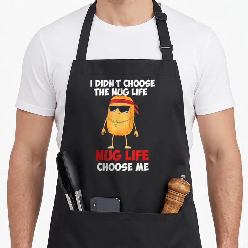 Funny nugget chicken print apron - Cartoon orange nugget with sunglasses and gold chain between white and red text — shown on a black cotton canvas apron against a white background