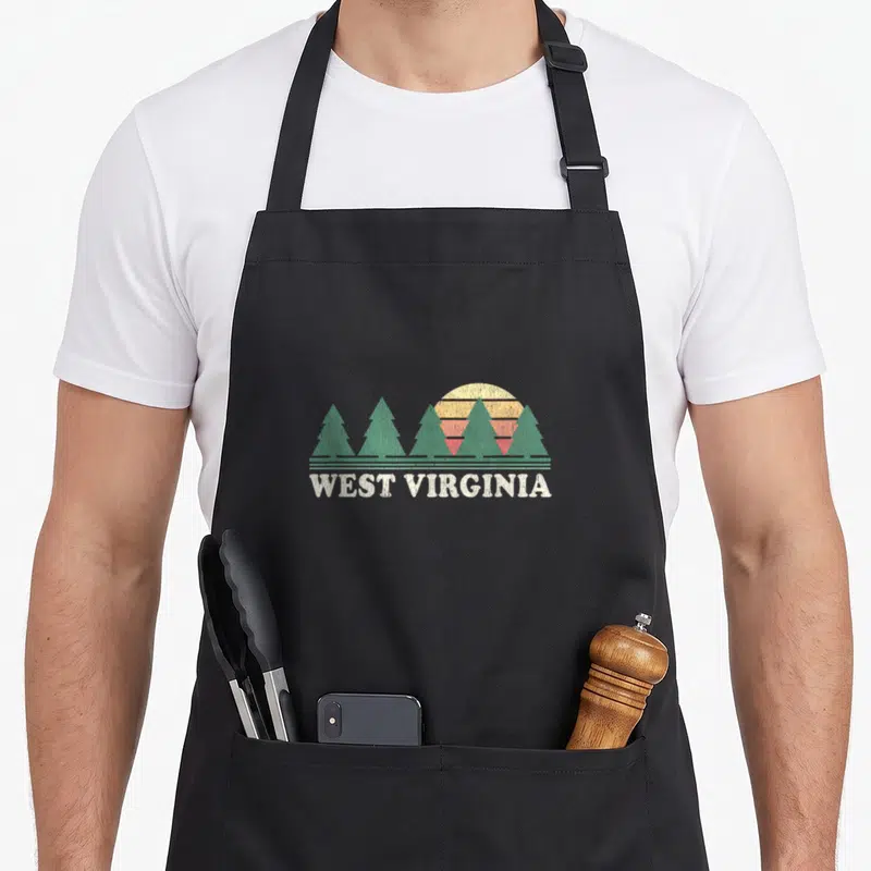 West 70s apron - Retro West Virginia text below green pine trees and striped sun in orange yellow off-white — shown on a black cotton canvas apron against a white background