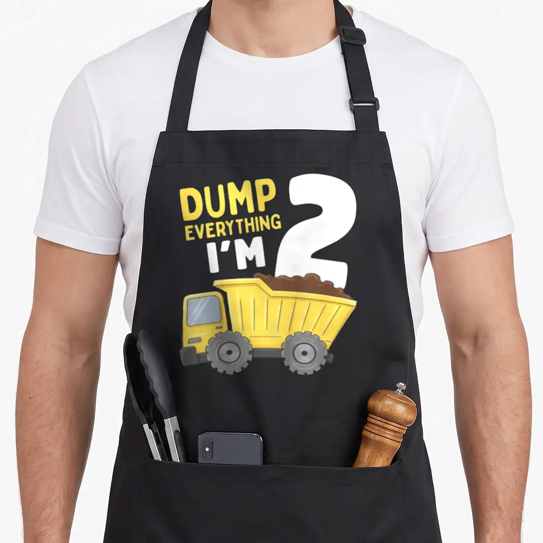 Construction 2nd truck apron - Yellow dump truck with brown dirt and text DUMP EVERYTHING I'M 2 in yellow and white — shown on a black cotton canvas apron against a white background