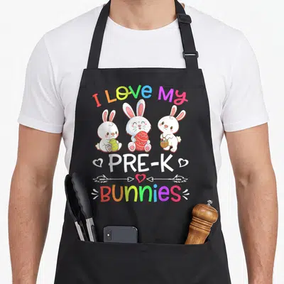 Pre teacher easter bunny aprons - Three cartoon bunnies holding eggs and baskets under rainbow text reading I Love My — shown on a black cotton canvas apron against a white background
