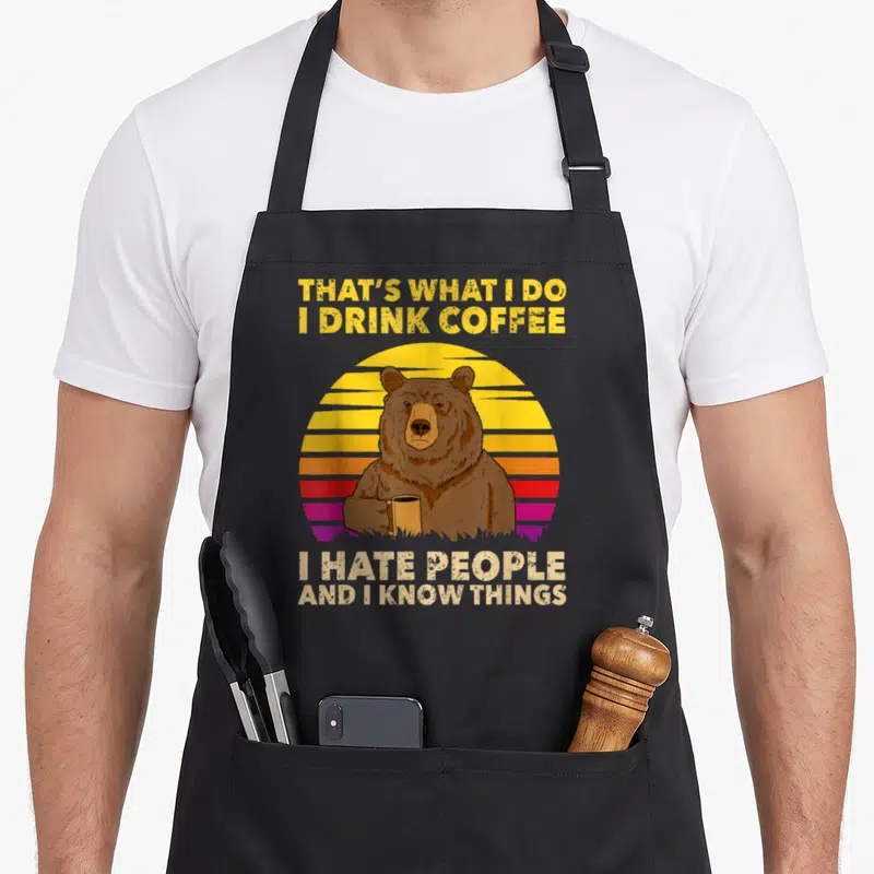 Drink hate bear coffee apron - Brown bear holding mug under yellow text and above white text on striped sunset — shown on a black cotton canvas apron against a white background