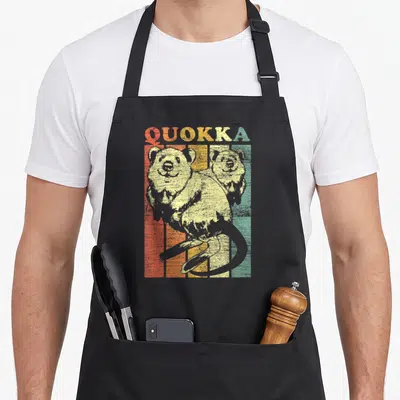 Quokka kangaroo apron - Two quokkas in front of vertical red orange yellow green beige stripes with QUOKKA text — shown on a black cotton canvas apron against a white background