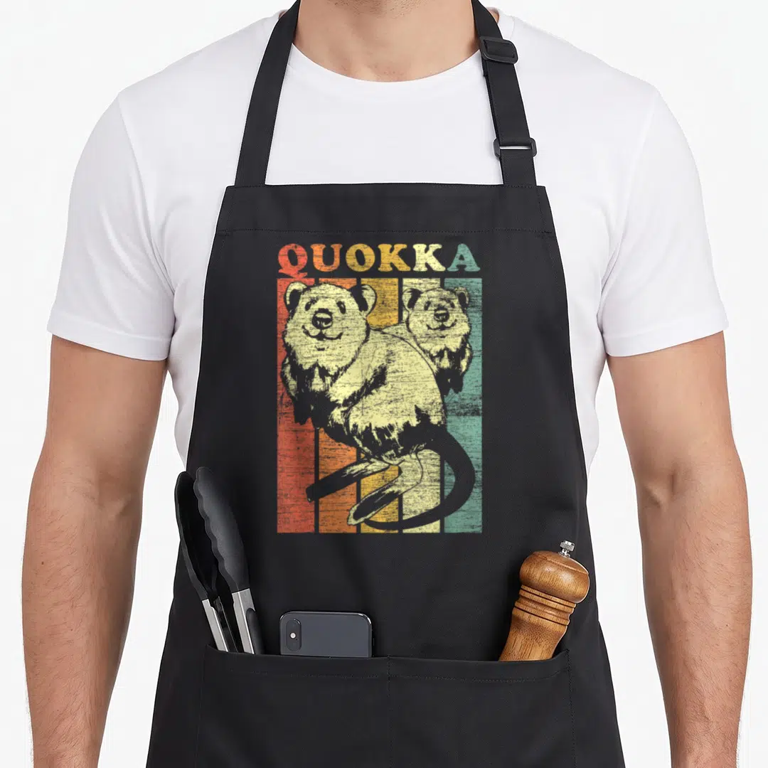 Quokka kangaroo apron - Two quokkas in front of vertical red orange yellow green beige stripes with QUOKKA text — shown on a black cotton canvas apron against a white background