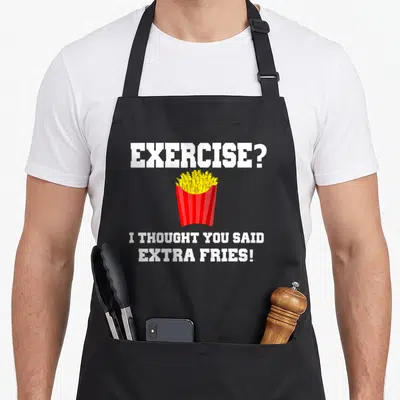 Fry an extra apron - White text reading exercise extra fries above a red container of yellow cartoon french fries — shown on a black cotton canvas apron against a white background