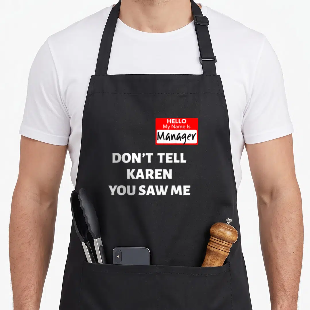 Manager halloween apron - Red name tag with Manager text above three lines of white Don't Tell Karen You Saw Me text — shown on a black cotton canvas apron against a white background