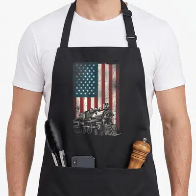 Steam american flag apron - White line art steam locomotive over distressed red white and blue USA flag — shown on a black cotton canvas apron against a white background