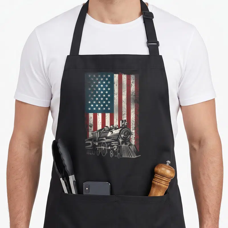 Steam american flag apron - White line art steam locomotive over distressed red white and blue USA flag — shown on a black cotton canvas apron against a white background