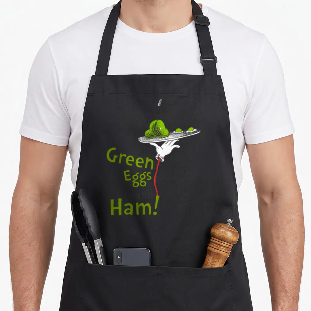Ham title egg apron - Cartoon hand holding green food plate with stacked Green Eggs and Ham text — shown on a black cotton canvas apron against a white background