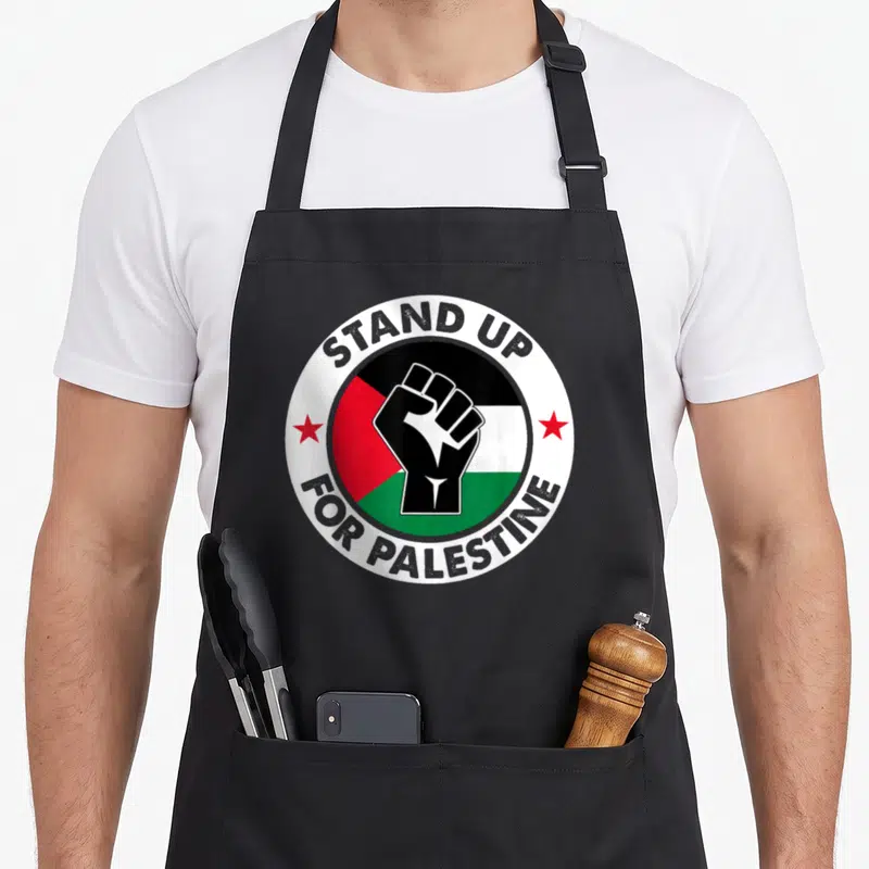 Free stand palestine apron - Raised fist on Palestinian flag colors with STAND UP FOR PALESTINE text and red stars — shown on a black cotton canvas apron against a white background