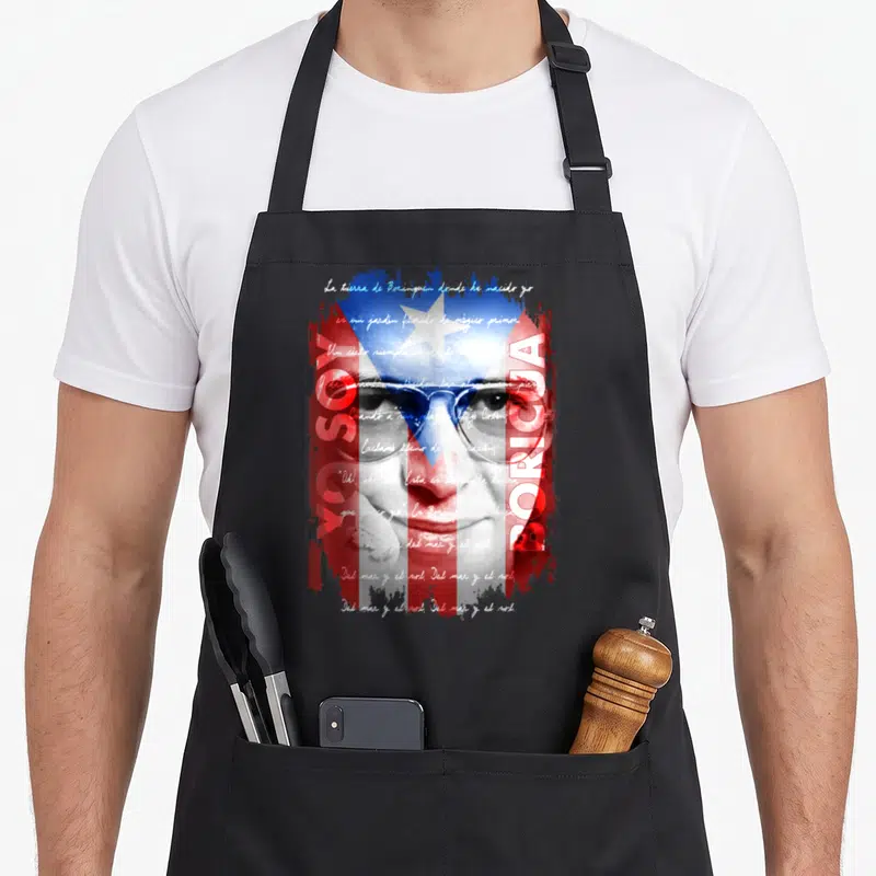 Hector soy apron - Woman face overlaid with Puerto Rican flag and Spanish lyrics in red white blue — shown on a black cotton canvas apron against a white background