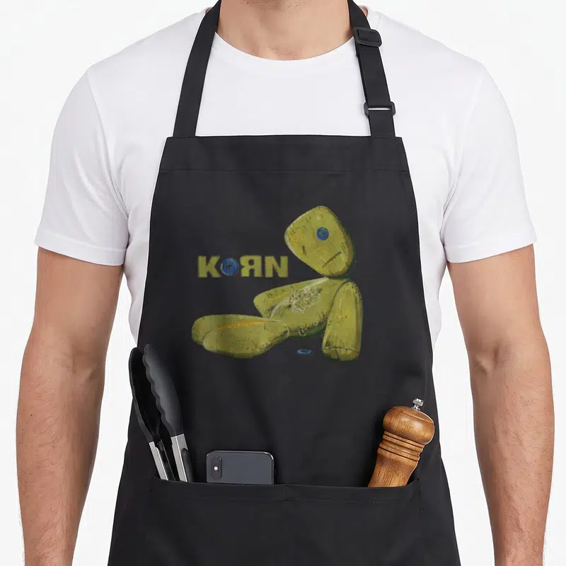 Korn doll apron - Olive green rag doll sitting next to KORN text with blue button replacing the O — shown on a black cotton canvas apron against a white background