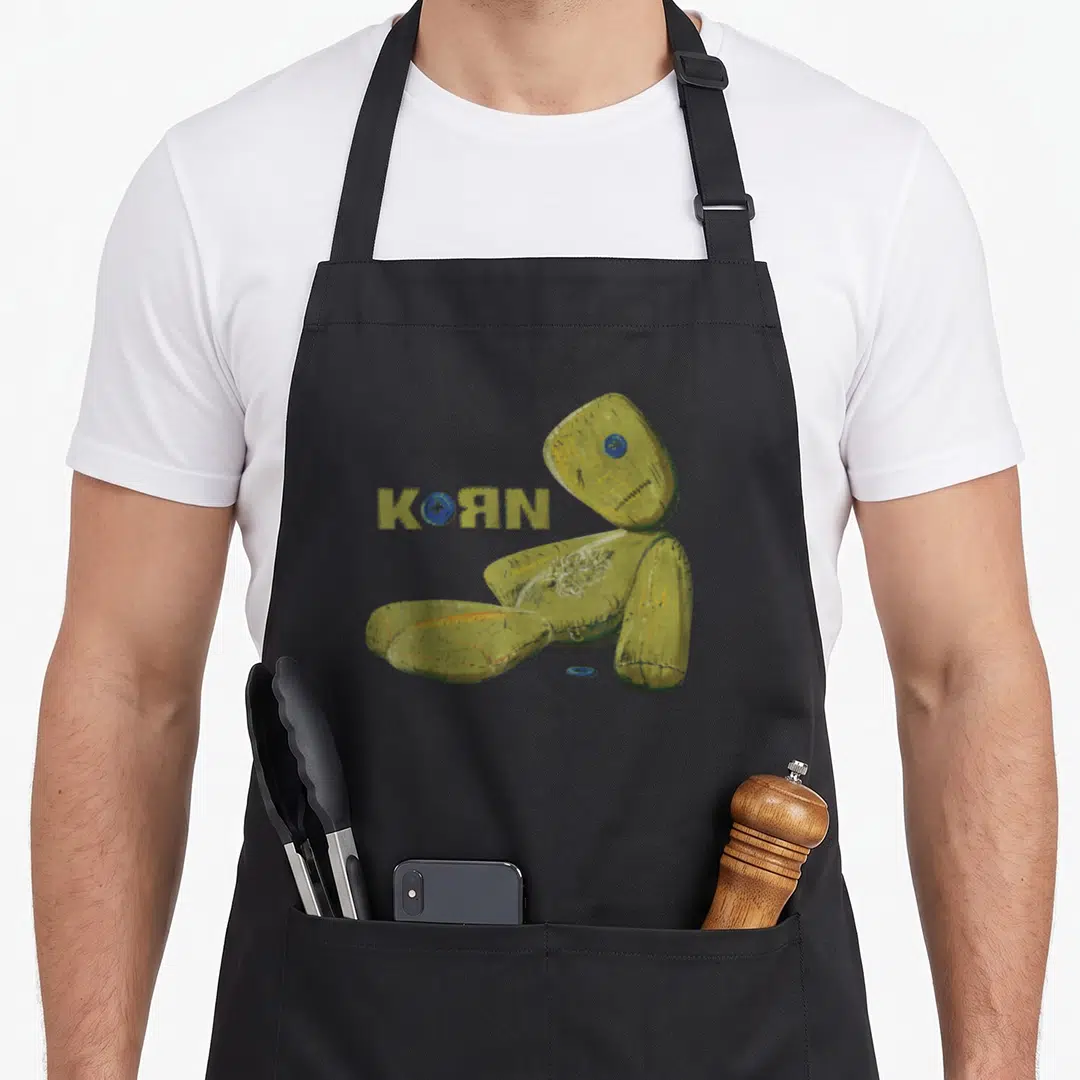 Korn doll apron - Olive green rag doll sitting next to KORN text with blue button replacing the O — shown on a black cotton canvas apron against a white background