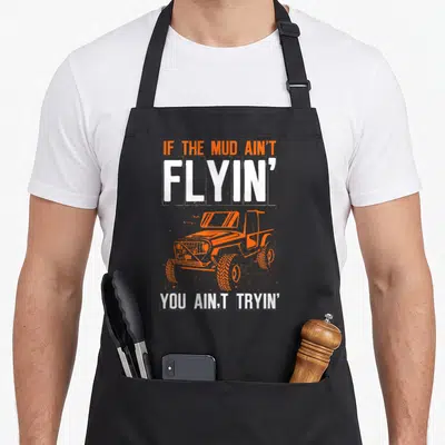 Mud road apron - Orange off-road vehicle between distressed white text blocks on black fabric — shown on a black cotton canvas apron against a white background