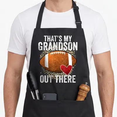 Grandson football grandpa grandma aprons - That's My Grandson Out There text above brown football and red heart — shown on a black cotton canvas apron against a white background
