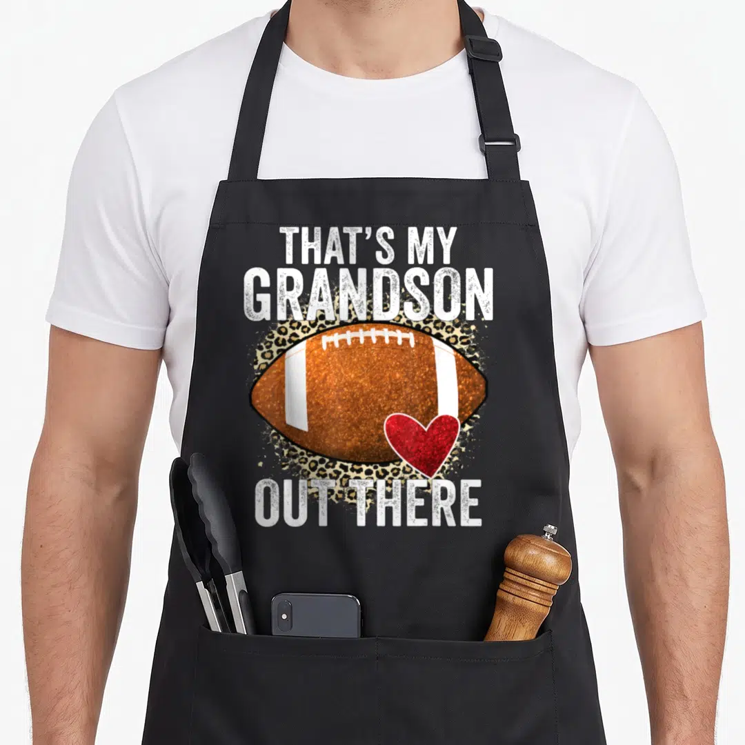 Grandson football grandpa grandma aprons - That's My Grandson Out There text above brown football and red heart — shown on a black cotton canvas apron against a white background