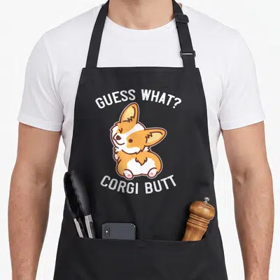 Buncho dog apron - Cartoon corgi in orange and white between GUESS WHAT? and CORGI BUTT text arcs — shown on a black cotton canvas apron against a white background