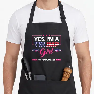 Apology novelty apron - Centered text YES IM A TRUMP Girl NO APOLOGIES with flag pattern and pink stars — shown on a black cotton canvas apron against a white background
