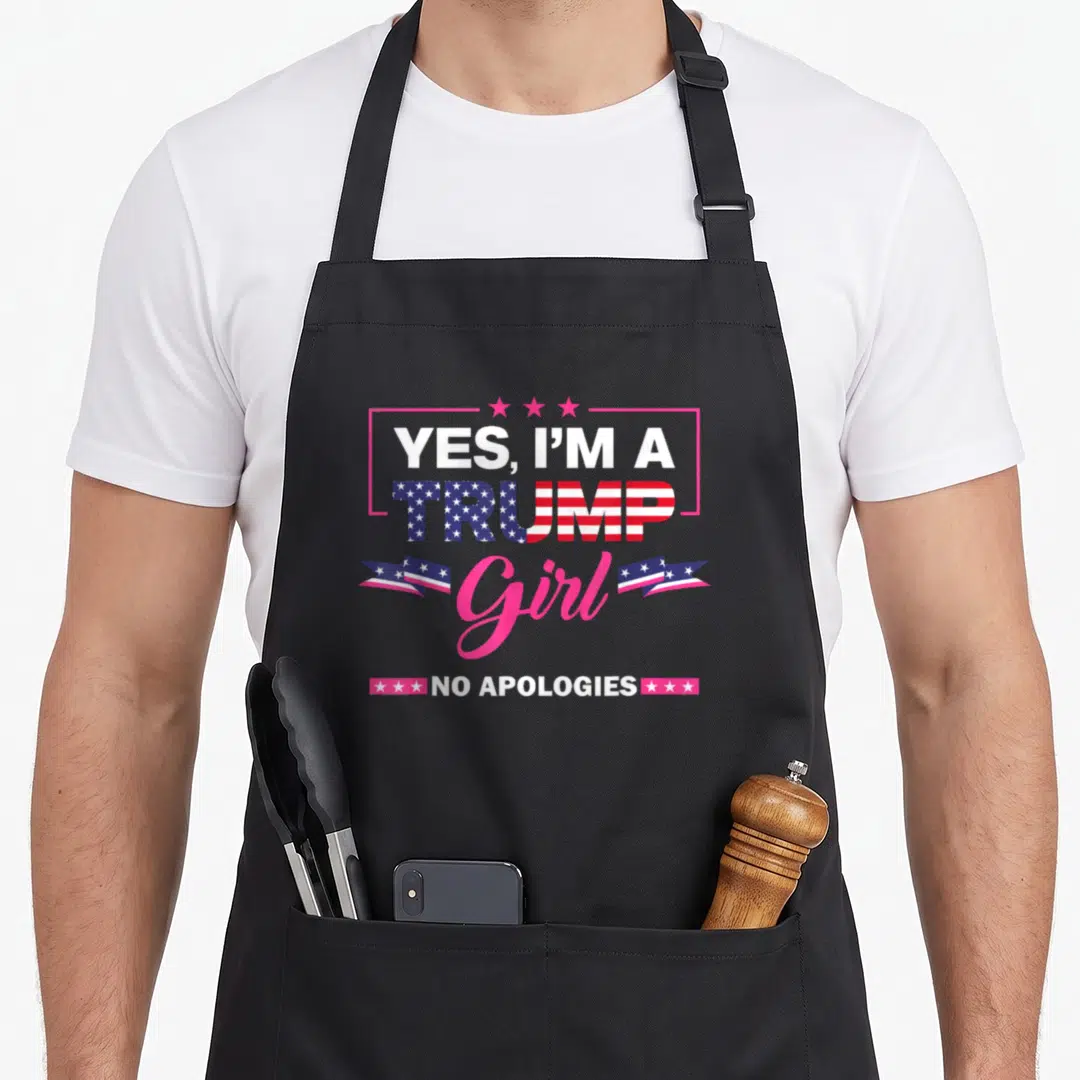 Apology novelty apron - Centered text YES IM A TRUMP Girl NO APOLOGIES with flag pattern and pink stars — shown on a black cotton canvas apron against a white background