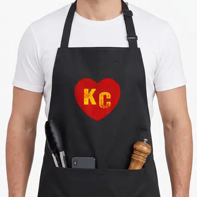 Funny kansas apron - Distressed red heart containing yellow KC letters centered on fabric — shown on a black cotton canvas apron against a white background
