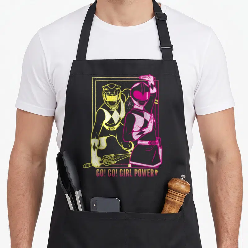 Cute rangers apron - Pink and yellow Power Rangers in a frame with GO! GO! GIRL POWER text below — shown on a black cotton canvas apron against a white background