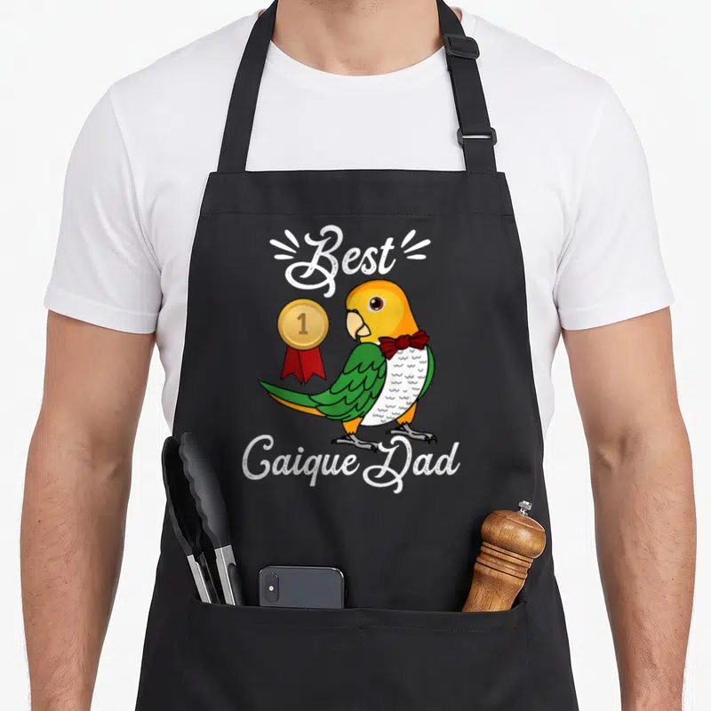 Bellied parrot dad apron - Cartoon caique parrot with gold medal and red bow tie, Best Caique Dad text — shown on a black cotton canvas apron against a white background