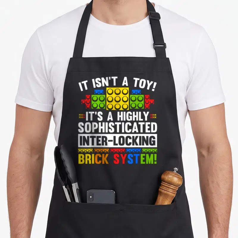 Brick grill master apron - Sarcasm text IT ISN'T A TOY above and below colorful interlocking toy bricks — shown on a black cotton canvas apron against a white background