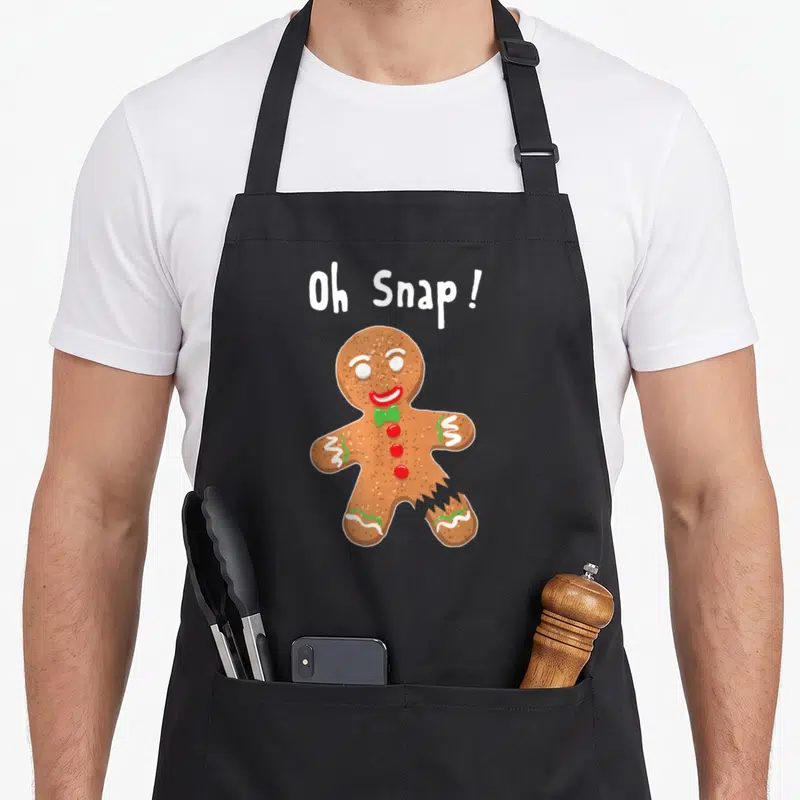 Gingerbread christmas cookie baking team aprons - Oh Snap text above broken gingerbread man with red buttons and green — shown on a black cotton canvas apron against a white background