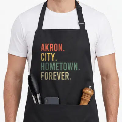 Akron forever home apron - Stacked text reading Akron City Hometown Forever in orange yellow green and tan — shown on a black cotton canvas apron against a white background