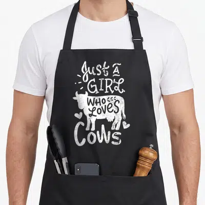 Cute cow apron - White distressed cow silhouette with stacked text and small hearts near legs — shown on a black cotton canvas apron against a white background