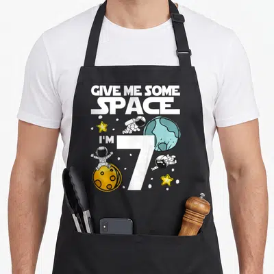 Planet birthday apron - Cartoon astronauts, blue planet, yellow moon, and text Give Me Some Space I'm 7 — shown on a black cotton canvas apron against a white background