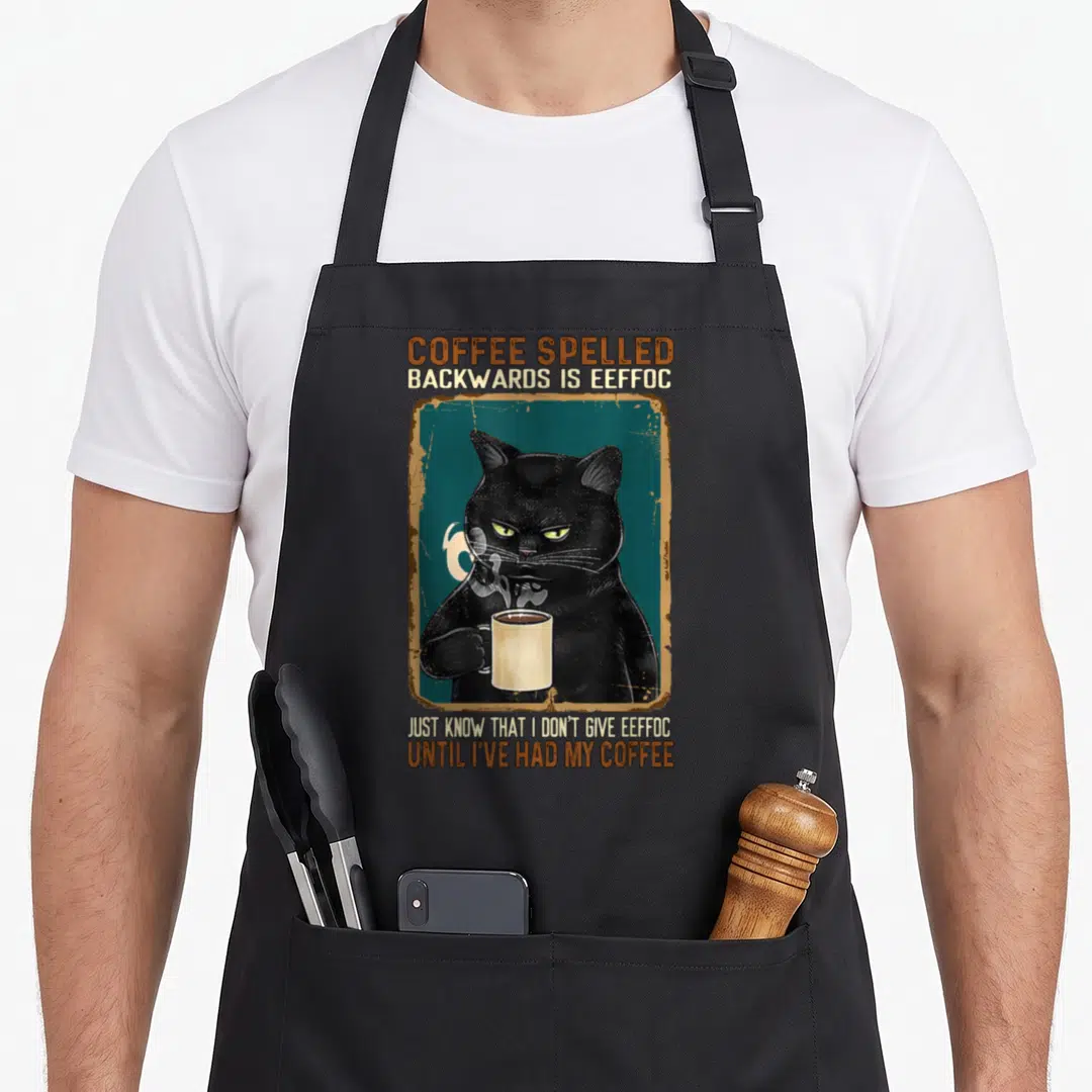 Spelled coffee aprons - Grumpy black cat holding mug with coffee spelled backwards text pun — shown on a black cotton canvas apron against a white background