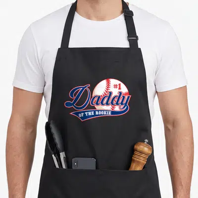Rookie daddy apron - #1 Daddy OF THE ROOKIE text over a baseball graphic in blue, red, and white — shown on a black cotton canvas apron against a white background